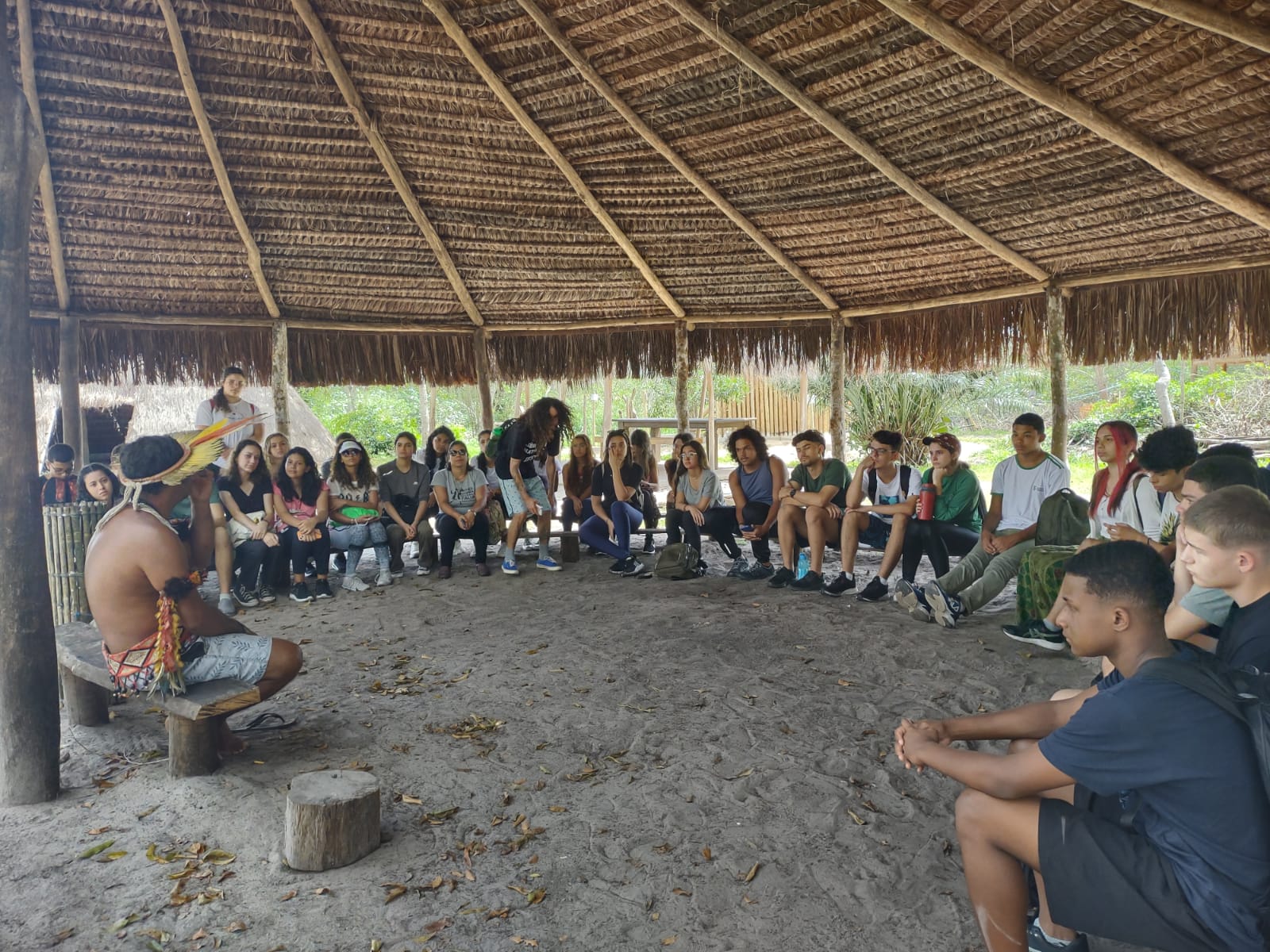 Estudantes e indígenas estão sentado e conversam em uma roda. Acima deles, há um telhado de palha.