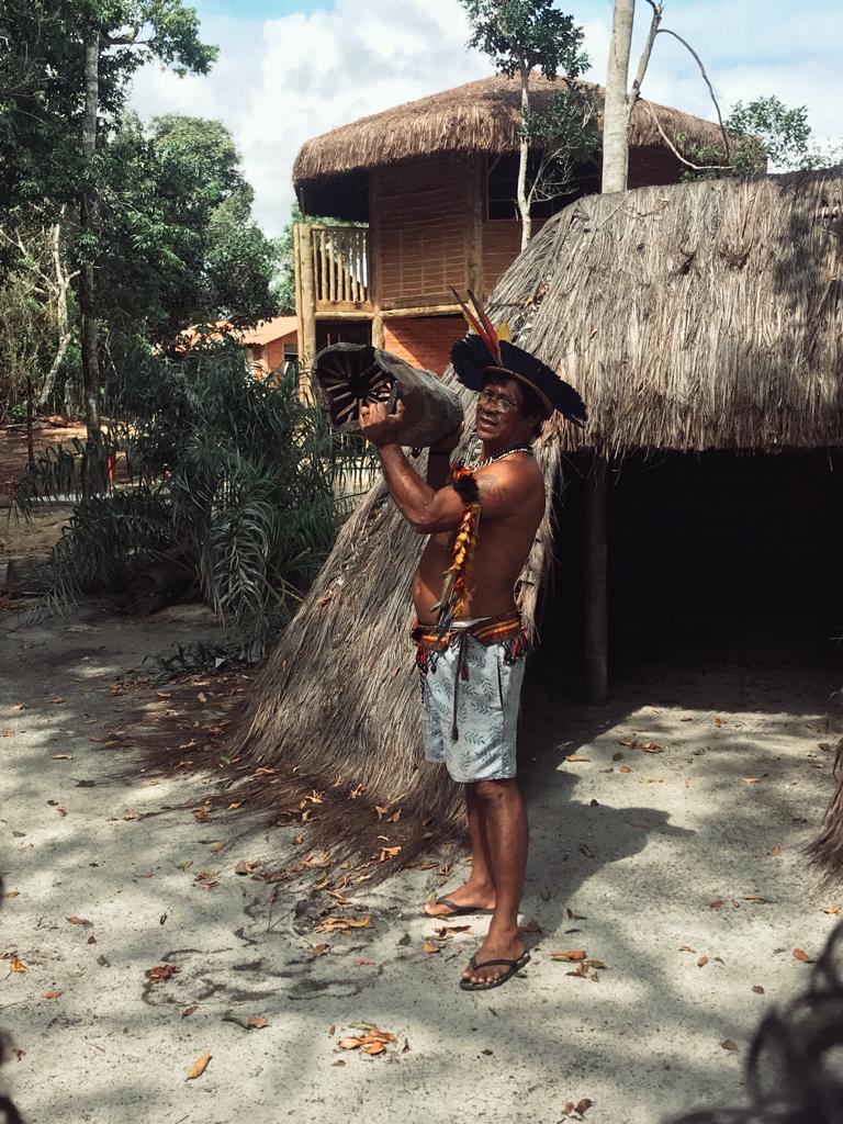 Um dos habitantes da Aldeia mostra uma tora de madeira em frente a uma oca.